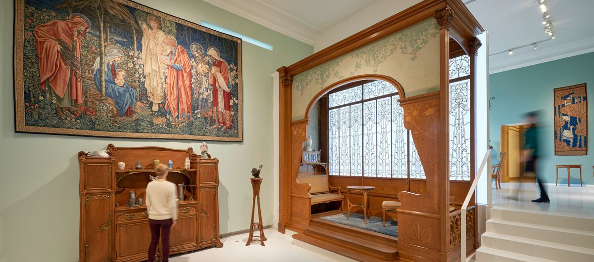 Foto eines Ausstellungsbereichs. Im Vordergrund an der Wand ein großer farbiger Wandteppich mit Szene der Geburt Jesu. Darunter eine Anrichte. Rechts daneben eine Holznische. Darin ein Sofa, runder Tisch und Stuhl. Die Möbel sind aus der Jugendstil-Zeit.