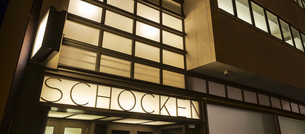 Foto einer mehrstöckigen Gebäudefront bei Nacht. Viele moderne Fenster zieren die Front. Im Inneren des Gebäudes brennt Licht. Über dem Eingang steht der Schriftzug "Schocken".