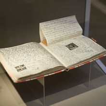 Foto einer originalgetreuen Kopie des Tagebuchs von Anne Frank. Das Buch liegt aufgeschlagen in einer Vitrine. Auf der linken Buchseite sind vier kleine Portraitfotos in den Text gelebt. Auf der rechten Seite ein größeres.