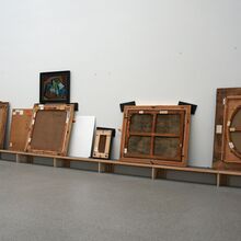 Foto von unterschiedlich großen Gemälden, die auf einer langen tiefen Holzbank an eine weiße Wand angelehnt stehen. Sie zeigen mit den Rückseiten zum Betrachtenden.