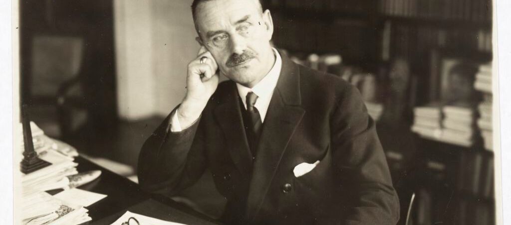 Schwarz-weiß Foto von Thomas Mann. Er sitzt mit einem Anzug bekleidet an einem Schreibtisch. Er stützt sich mit seinem rechten Ellenbogen auf den Schreibtisch, lehnt seinen Kopf an seine rechte Hand und schaut nach links. Vor ihm Papierstapel.