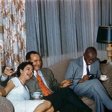 Foto, das wie ein privater Schnappschuss wirkt. Auf einem Sofa sitzen eine Frau und zwei Männer, die befreit lachen. Alle haben eine Zigarette in der Hand. Die Frau und ein Mann haben eine Kaffeetasse in der Hand, der andere Mann ein Schnapsglas.