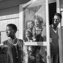 Schwarz-weiß Foto in Nahaufnahme vor einem Haus. Zwei Jugendliche stehen vor der Haustür. Eine Frau steht zwischen Haustür und Fliegengitter und schaut heraus. Drei Jugendliche stehen hinter dem Fliegengitter. Alle Menschen haben eine dunkele Haut.