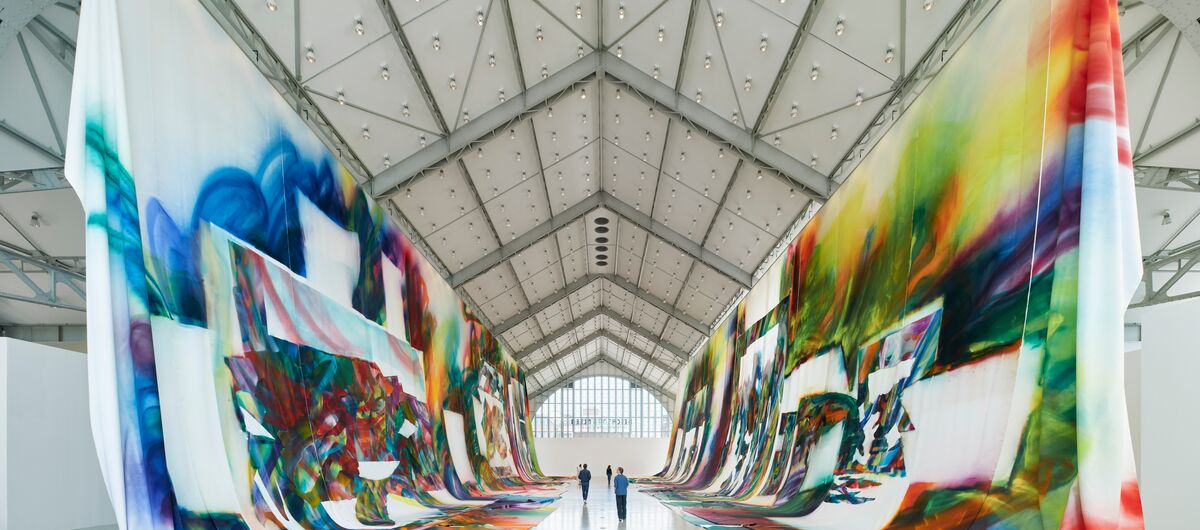 Foto der Innenansicht der Deichtorhalle. rechts und links hängen von den Decken lange weiße Stoffbahnen, die teils auch den Boden bedecken. Auf dem Stoff sind bunte, abstrakte graffitiartige Darstellungen, die durch weiße Bereiche unterbrochen werden.