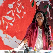 Das Foto zeigt die Künstlerin Portia Zvavahera in ihrem Studio. Sie ist eine schwarze Frau mit Dreadlocks bis zum Bauch. Ihr Blick ist freundlich. Sie stützt sich an ihrem Tisch ab, auf denen Pinsel und Farbdosen stehen. Im Hintergrund ein Gemälde.