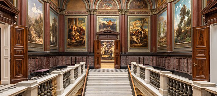Foto des Treppenhauses in der Lichtwark-Galerie der Hamburger Kunsthalle. Eine Steintreppe, die in der unteren Bildmitte ist, führt in ein weiteres Geschoss mit historischen Bildern und Wandverzierungen.