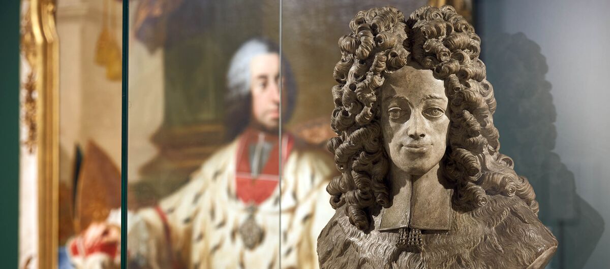 Foto einer Vitrine, in der eine Büste aus dem Zeitalter des Barock steht. Im Hintergrund Ist ein großes buntes Gemälde zu sehen, auf dem ein sitzender Mann ebenfalls aus der Zeit des Barock abgebildet ist.