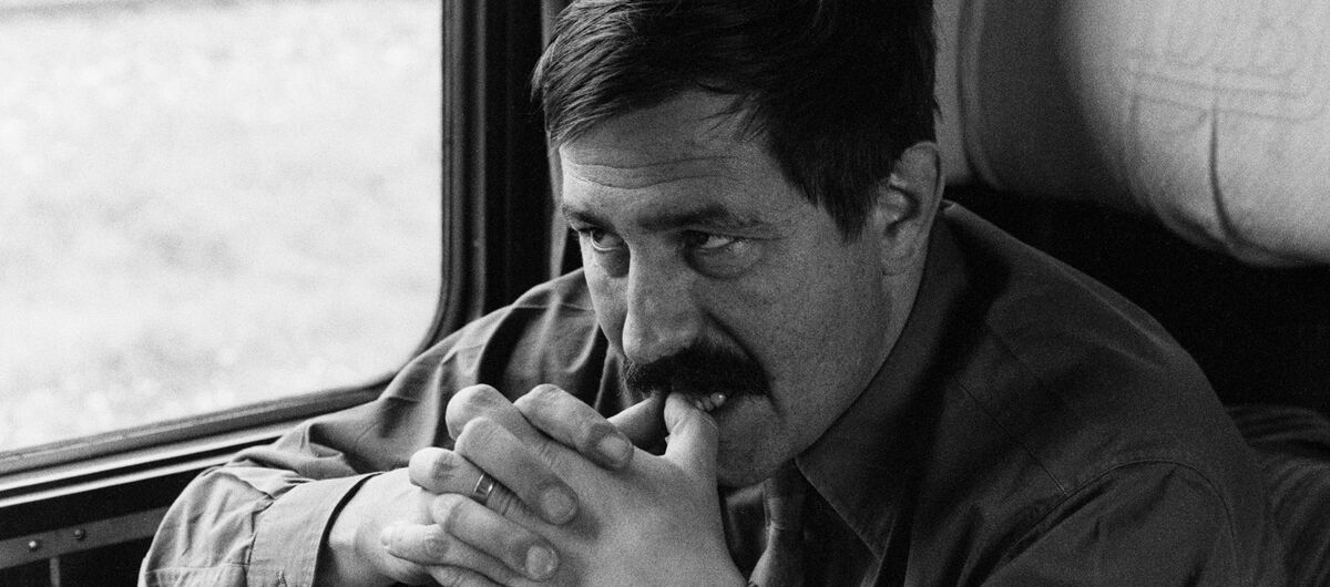Schwarz-weiß Foto von Günter Grass in einem Zugabteil. Er sitzt am Fenster und schaut auf den gegenüberliegenden Sitz. Sein rechtes Bein ist aufgestellt, die Ellenbogen auf die Knie gestützt, die Hände gefaltet und die Daumenspitzen an die Zähne gelegt.