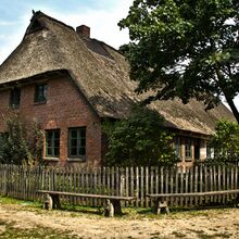 Foto eines geklinkerten Bauernhauses mit Reetdach und grünen Sprossenfenstern. Das Haus ist von einem Holzzaun eingefasst. Vor diesem stehen zwei einfache Holzbänke. Im Garten des Hauses stehen ein großer Baum und weitere Büsche.