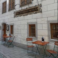 Foto eines Gebäudes. Davor stehen zwei kleine Klapptische mit je zwei Klappstühlen. An dem Haus ist ein Schild befestigt, auf dem "Bier Oktoberfest Museum" steht.
