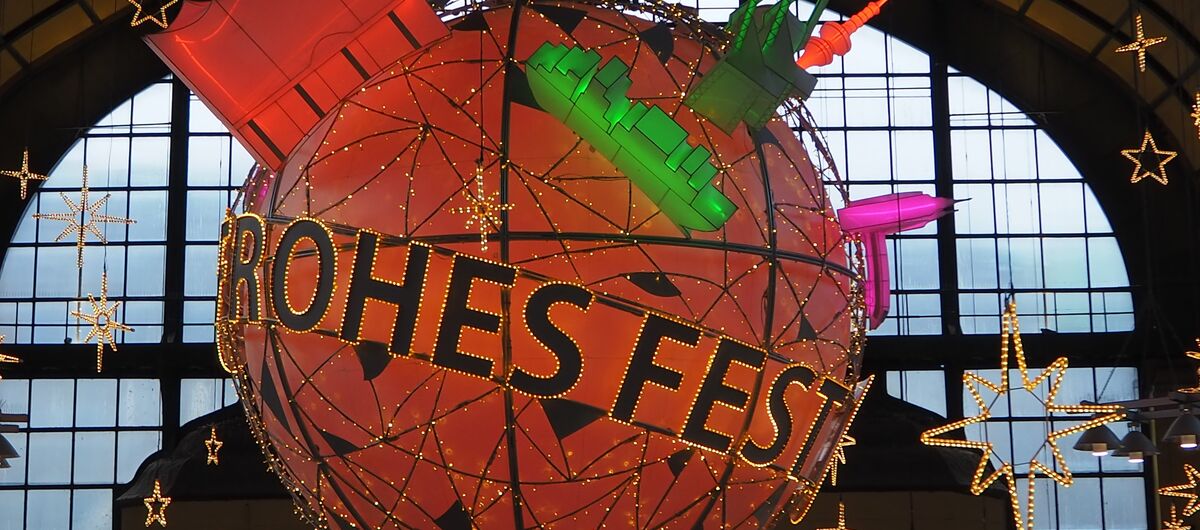 Foto von Weihnachtsbeleuchtung im Hamburger Hauptbahnhof: Eine rote Kugel mit dem Schriftzug Frohes Fest, die ca. zwei Meter im Durchmesser ist, hängt unter der Decke. Darauf sind weitere unterschiedliche Symbole Hamburgs.