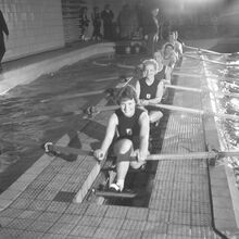Schwarz-weiß Foto mit dem Titel „Versehrten-Rudern“, aufgenommen am 19.2.1962 in Berlin. In einem festinstallierte Rudertrainingsgerät mit sieben Plätzen sitzen in Sportkleidung freundlich schauende Frauen und Männer.