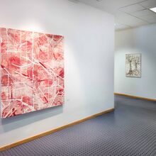 Foto eines Flures mit blauem Teppichboden und weißen Wänden. An der Wand im rechten Vordergrund hängt eine große quadratische Leinwand mit abstrakter Malerei mit roten, braunen und weißen Farben. Im Hintergrund hängt ein kleineres Bild mit zwei Bäumen.