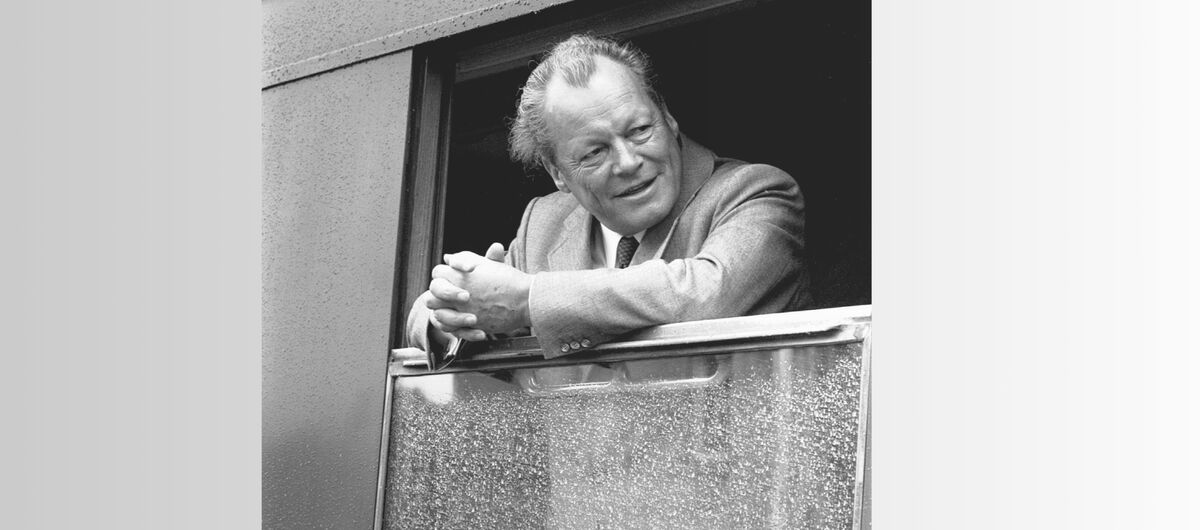 Schwarz-weiß Foto von Willy Brandt, der in einem Zug steht und sich mit seinen gefalteten Händen auf das heruntergelassene Fenster abstützt. Er schaut freundlich nach links. Das Fenster ist nass.
