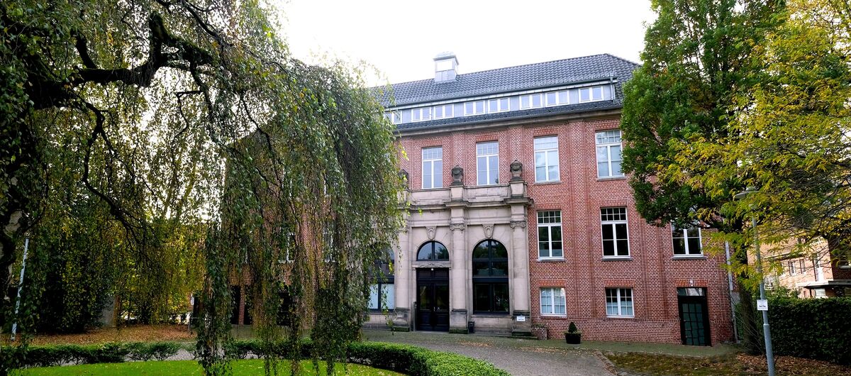 Historisches Backsteingebäude mit großen Fenstern und einem klassischen Eingangsbereich mit Säulen. Im Vordergrund befindet sich eine gepflegte Gartenanlage mit einem Kopfsteinpflasterweg, gesäumt von grünen Hecken und Bäumen.