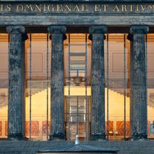 Foto des Eingangsbereichs des Alten Museums in Berlin. Treppen führen hoch zu einer großen Glasfront mit Glastür, die durch sechs pompöse Säulen geschützt wird. Darüber eine lateinische Inschrift in goldenen Buchstaben. Im Inneren ist ein warmes Licht.