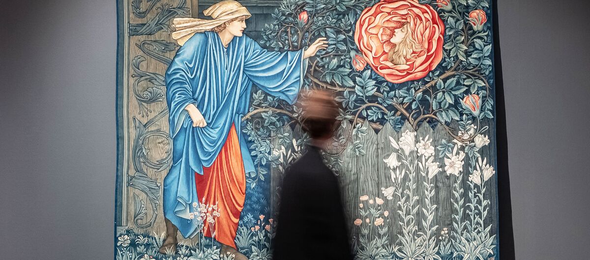 Foto eines angeleuchteten Teppichs vor einer grauen Wand, vor dem ein verschwommener Mensch herläuft. Auf dem Teppich ist ein Mann im Garten abgebildet. Er streckt seinen linken Arm zu einer überdimensionierten Rose mit Frauenkopf.