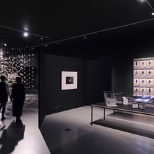 Schwarz-weiß Foto eines Ausstellungsbereichs. Es liegen Exponate auf zwei Tischen. An den beiden schwarzen Wänden drumherum sind ein einzelnes sowie eine Fotoreihe mit über 24 Fotos zu sehen. Viele kleine Zettel hängen links von der Decke.