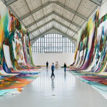 Foto der Innenansicht der Deichtorhalle. rechts und links hängen von den Decken lange weiße Stoffbahnen, die teils auch den Boden bedecken. Auf dem Stoff sind bunte, abstrakte graffitiartige Darstellungen, die durch weiße Bereiche unterbrochen werden.