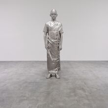 Foto einer Statue, die vor einer weißen Wand und auf grauem Betonboden steht. Sie ist silbern. Dargestellt wird ein geradestehender Mensch mit kurzen Haaren, knöchellanger Tunika und Schwert in seiner rechten Hand. Der Gesichtsausdruck ist neutral.