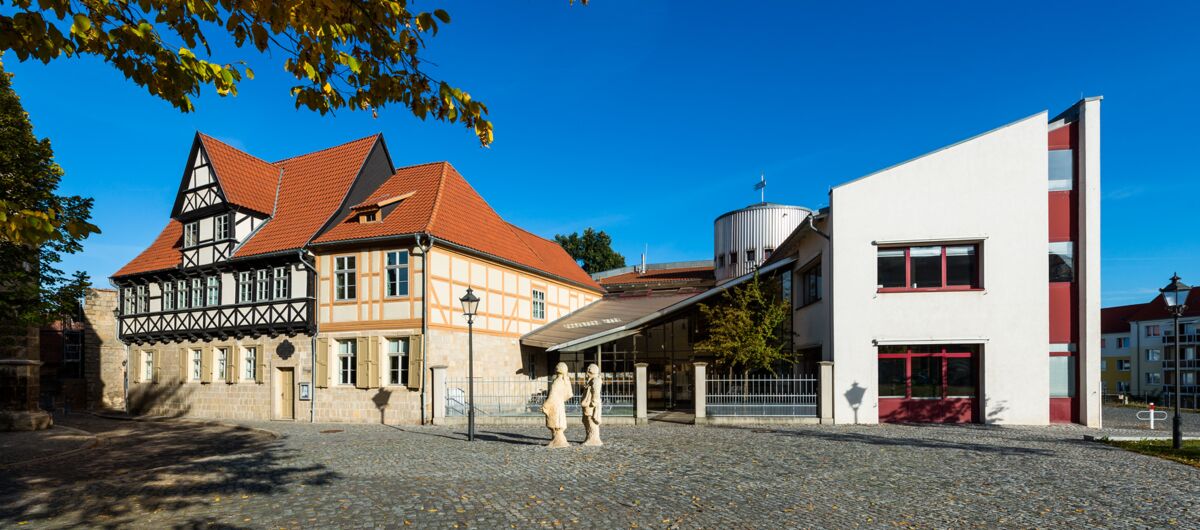 Zwei Gebäude nebeneinander, links ein dreigeschossiges Fachwerkhaus mit hellem Stein, dunklem Gebälk und rotem Ziegelspitzdach. Rechts ein Neubau. Verbunden werden beide Gebäude mit einem schrägen Vordach, in dessen Mitte ein runder Turm steht.