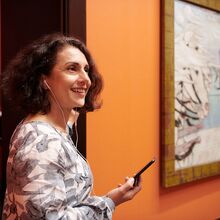 Foto einer lächelnden Frau, deren Blick und Körperausrichtung nach rechts orientiert ist, in einer Kunstausstellung. Sie hat ein Handy in ihrer rechten Hand, das sie vor sich hält und mit Kopfhörern verbunden sind, die in ihren Ohren stecken.