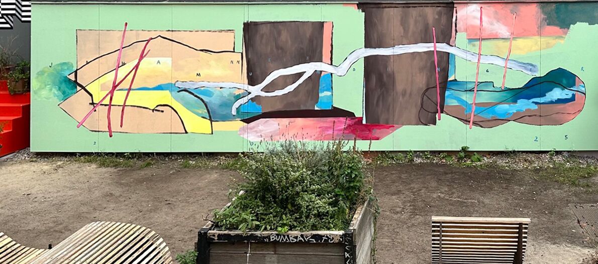 Foto einer abstrakt bemalten Wand, die ca. 8 Meter lang und 2,5 Meter hoch ist. Davor stehen ein bepflanztes Hochbeet sowie rechts und links davon zwei Holzliegen. Der Boden besteht aus Sand und Erde.
