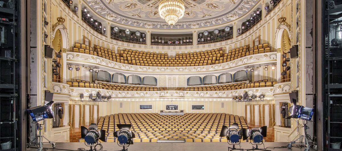 Foto von der Bühne aus in den prachtvollen Zuschauersaal des Staatsschauspiel Dresden. Im Vordergrund stehen auf der Bühne Scheinwerfer, die auf den Saal gerichtet sind. Der Saal ist in Creme- und Goldfarben gehalten.