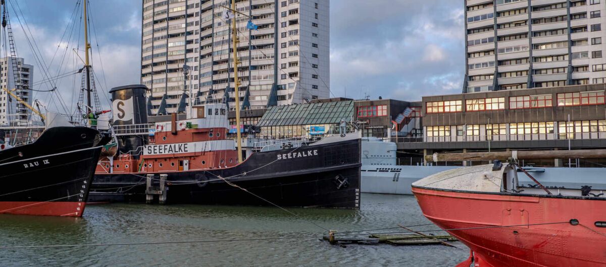Hafenszenerie mit 3 Schiffen, die im Wasser dümpeln. Die Seefalke liegt mittig und hat sowohl Segelaufbauten, als auch einen Dampfer-Schornstein an Deck.