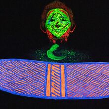 Das Bild zeigt im Vordergrund eine Art Schild, in das kunstvoll ein systematisches Muster aus Strichen geschnitzt ist - mit blauer und orangener Farbe bemalt. Dahinter ein Mensch, dessen Gesicht und Brust mit fluoreszierender Farbe bemalt wurde.