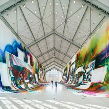 Foto der Innenansicht der Deichtorhalle. rechts und links hängen von den Decken lange weiße Stoffbahnen, die teils auch den Boden bedecken. Auf dem Stoff sind bunte, abstrakte graffitiartige Darstellungen, die durch weiße Bereiche unterbrochen werden.