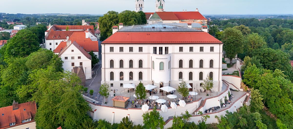 Luftfoto des Diözesanmuseum Freising. Auf einem Berg steht das viergeschossige Haus. Davor ist ein Platz, auf dem Sonnenschirme stehen und der halbrund eingemauert ist. grüne Bäume schmiegen sich dicht hangabwärts an diesen Komplex.