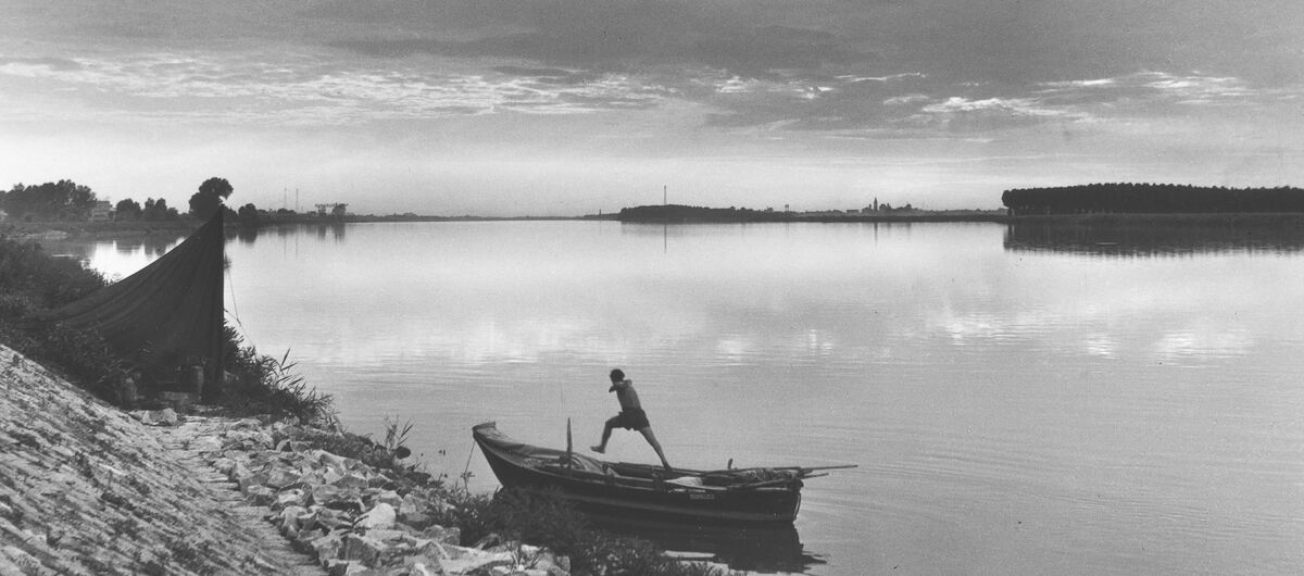 Schwarz-weiß Foto, auf dem ein einfaches kleines Holzboot am steinigen Rande eines stillen Sees liegt. Von dem Boot scheint ein Mensch herunterzuspringen. Der Blick über den See ist weit.