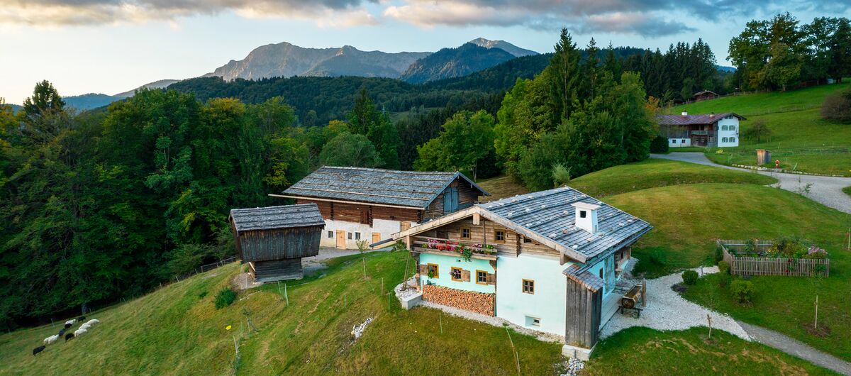 Luftaufnahme eines Bereichs des Freilichtmuseums Glentleiten. Traditionelle Hofgebäude stehen auf einem Berg. Auf der Abschüssigen Wiese unter den Gebäuden stehen weiße und schwarze Schafe. Im Hintergrund sind Wald und Berge zu sehen.