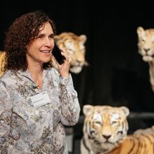 Foto einer Frau mittleren Alters, die ein Namensschild an der Bluse trägt und ein Handy an ihr linkes Ohr hält. Sie schaut freundlich. Im Hintergrund sind drei Tiger-Figuren vor einem schwarzen Hintergrund zu sehen.