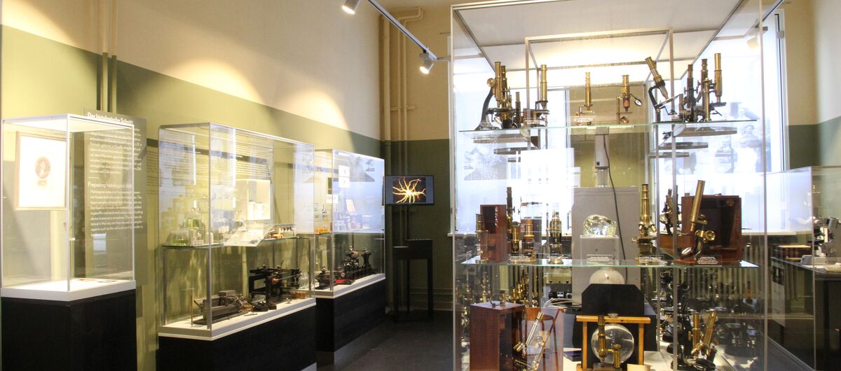 Foto eines Ausstellungsbereichs. In einem kleinen Raum mit Fenster an der Stirnseite stehen schmale Vitrinen an den Wänden und eine große Vitrine in der Mitte. In der mittigen sind historische Mikroskope zu sehen, in den anderen weitere technische Geräte.