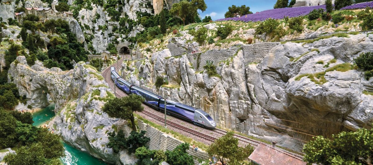 Modell einer bergigen Felslandschaft, durch die eine Eisenbahntrasse führt. Ein Fluss schlängelt sich türkisblau neben der Schiene. Auf dieser der französische Hochgeschwindigkeitszug TGV. Lila Lavendelfelder im Hintergrund.