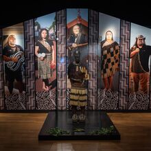 Foto einer Ausstellungsfläche. Der Hintergrund ist schwarz, davor stehen angestrahlte Ganzkörperfotos von Einwohnern Neuseelands. Vor dieser Fotowand liegt eine schwarze Platte, auf der eine dunkelhäutig Figur mit ursprünglicher Kleidung steht.