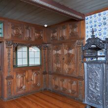 Foto einer barocken Bauernstube. Der Boden ist ebenso wie die kunstvoll geschnitzte Tür und die Wände links und mittig im Foto aus dunklem Holz. Die rechte Fotoseite wird durch eine mit blau-weißen kleinen Fliesen und einem kunstvollen Metallofen geprägt.