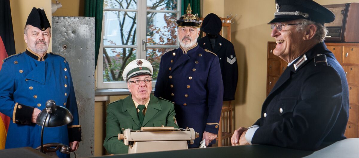 Vier Männer in einem Raum, drei stehend, einer sitzend. Alle tragen unterschiedliche Uniformen, die die verschiedenen Epochen der Polizeikleidung darstellen. Auf ihren Köpfen Hüte - von Pickelhaube über Barett bis zu Schiebermützen ist alles dabei.
