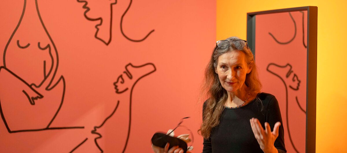 Eine Kulturvermittlerin vor einer orange-roten Wand mit verschiedenen skizzierten Körpern, im Hintergrund ein Spiegel.