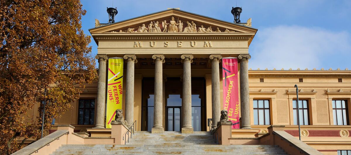 Foto vom prächtigen Eingangsbereichs des Staatlichen Museums Schwerin. Das Gebäude ist sandsteinfarbig. Im Vordergrund führt eine lange breite Treppe zu sechs Säulen, hinter denen sich die Eingangstür befindet.