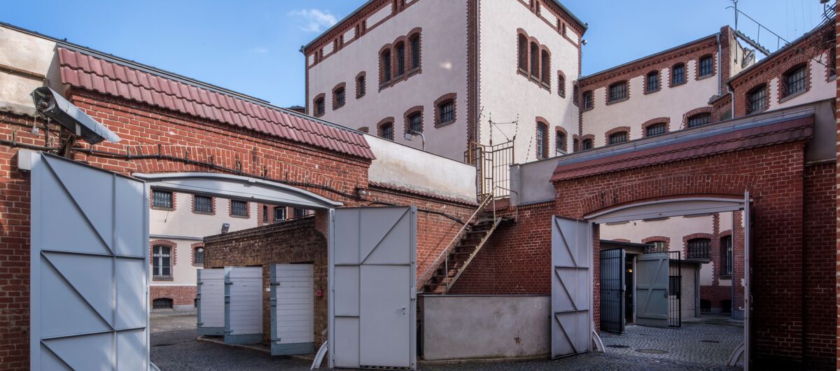 Außenansicht der Gedenkstätte Lindenstraße. Das verschachtelte Backsteingebäude ist teils weiß verputzt. Der Boden ist aus Kopfsteinpflaster. Im Vordergrund sind rechts und links zwei Durchgäng mit Flügeltüren zu sehen, die zu dem Hauptgebäude führen.