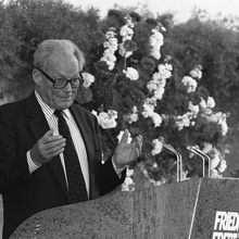 Schwarz-weiß Foto von Willy Brandt im Anzug während einer Rede. Er steht an einem Pult mit schwarzer Brille, Blick in Richtung Zuhörende und Händen auf Brusthöhe. Hinter ihm eine Steinwand an der Hecke und Blumen hinabranken.