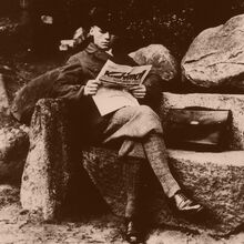 Schwarz-weiß Foto eines jungen gut gekleideten Mannes, der mit überschlagenen Beinen auf einer Bank aus großen Steinen sitzt und Zeitung liest. Neben ihm auf der Bank liegt eine Aktentasche.