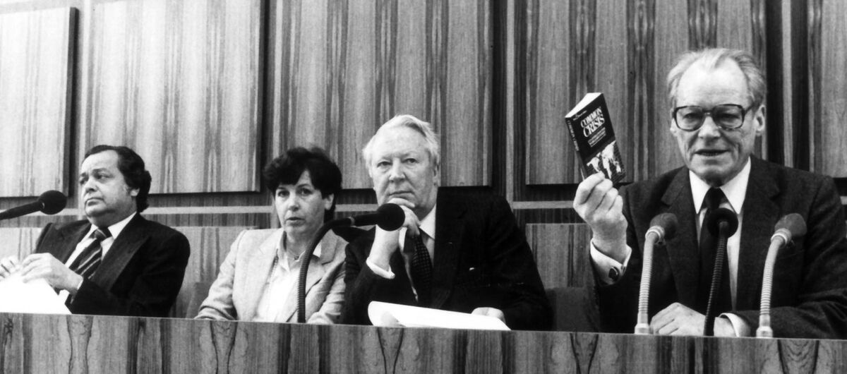 Schwarz-weiß Foto von einer Frau und drei Männern, die Nebeneinander hinter einem Pult mit Mikrofonen sitzen. ganz rechts ist Willy Brandt, der in drei Mikrofone spricht und mit seiner rechten Hand ein Buch hochhält.