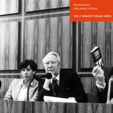 Schwarz-weiß Foto von einer Frau und drei Männern, die Nebeneinander hinter einem Pult mit Mikrofonen sitzen. ganz rechts ist Willy Brandt, der in drei Mikrofone spricht und mit seiner rechten Hand ein Buch hochhält.