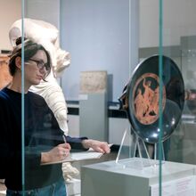 Foto eines Ausstellungsbereichs. Vor einer Glasvitrine, in der eine antike Schale ausgestellt ist, steht eine Frau mit Stift in ihrer rechten Hand, die auf einem dünnen Brett mit Papier liegt. Sie betrachtet das ausgestellte Objekt.