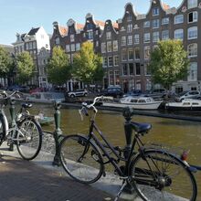 Foto einer Gracht. Im Vordergrund stehen zwei Fahrräder angelehnt an einem Geländer. Auf der gegenüberliegenden Seite sind Boote festgemacht. Dahinter ist eine Hausreihe.