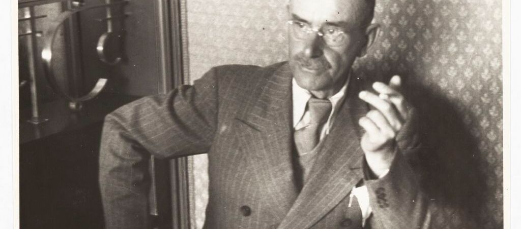 Schwarz-weiß Foto von Thomas Mann. Er sitzt mit einem Anzug bekleidet an einem Tisch. Er stützt sich mit seinem linken Ellenbogen auf diesen und hält in der linken Hand eine Zigarette. Er schaut nach rechts. Vor ihm Gläser und Geschirr.